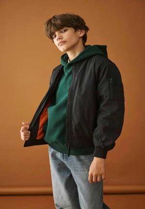 Jauns cilvēks zaļā kapučjakā un melnā bombers jakā, tur jakas atloku vaļā, lai parādītu oranžo oderi, stāvot pretī brūnam fonam.