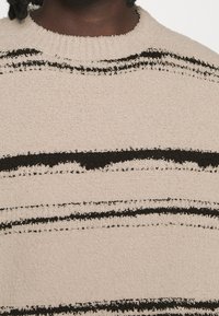 Detailní záběr na béžový texturovaný svetr s nepravidelnými černými vodorovnými pruhy, který nosí osoba s tmavou pletí a spletenými vlasy.