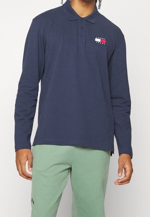 Osoba nosząca granatową, długorękawową koszulkę polo z logo Tommy Jeans i jasnozielone spodnie, stojąca na jednolitym tle.