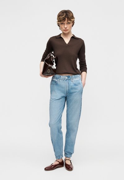 Γυναίκα που φοράει μακρυμάνικη μπλούζα σκούρου καφέ χρώματος, ανοιχτόχρωμα τζιν με μανσέτες, καφέ φλατ παπούτσια και κρατάει μια τσάντα ώμου σκούρου καφέ χρώματος.