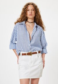 Blauw-witte gestreepte overhemd met opgerolde mouwen, gecombineerd met een witte denim rok en een bruine riem met een metalen gesp.