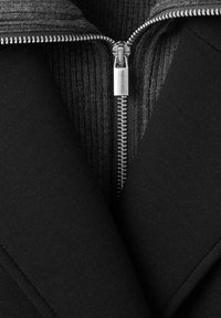 Chaqueta negra con cuello acanalado gris, con un cierre de metal que va por el frente, con una textura suave y un diseño entallado.