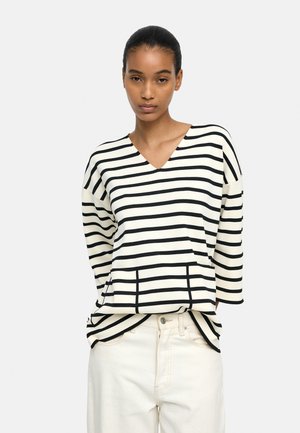 Młoda kobieta ubrana w czarno-biały poziomo paskowany sweter z dekoltem w serek oraz białe spodnie, stojąca na jednolitym tle.