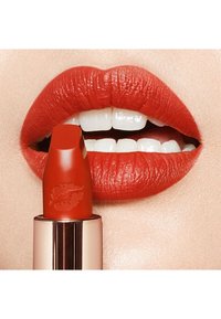 Red lipstick tube partially visible with a lipstick bullet resembling a kiss mark, applied to smooth, bright red lips over white teeth.