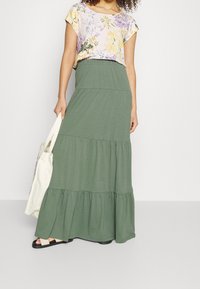 Wielowarstwowa, zielona maxi spódnica wykonana z miękkiego materiału; posiada elastyczną talię i luźny fason. Połączona z kwiecistym topem z krótkim rękawem.