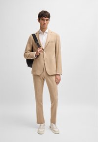 Costume beige avec un blazer à simple boutonnage et un pantalon à plat. Tissu texturé, associé à une chemise blanche et des baskets. Sac noir sur l'épaule.