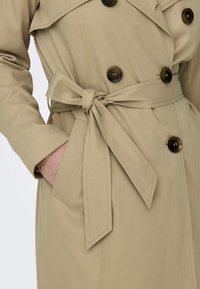 Trench-coat beige avec un design croisé, de gros boutons, une taille cintrée avec une ceinture et des poches latérales. Texture de tissu lisse.