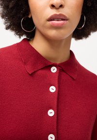 Pulôver de malha vermelho com decote colarinho. Apresenta quatro fechos de botão brancos e uma textura suave. Acento de brincos em argola visíveis.