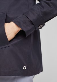 Morska niebieska wodoodporna kurtka z rękawem detalem w postaci guzika i widocznym szwem, z boczną kieszenią oraz okrągłym metalowym akcentem w pobliżu dolnej krawędzi.