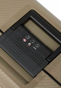 Valigia rigida con superficie beige testurizzata. Presenta un meccanismo di chiusura nero con tre pulsanti circolari e un foro per la chiave visibile.