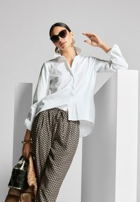 Witte button-up shirt, patroon bruine broek, zwarte handtas, oversized zonnebril en gouden creolen. Glad, gestructureerd materiaal en een relaxed fit.
