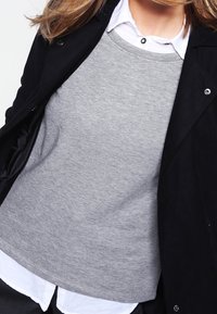 Kobieta ubrana w warstwowy strój, z białą koszulą z kołnierzykiem pod szarym swetrem i czarną kurtką na wierzchu.