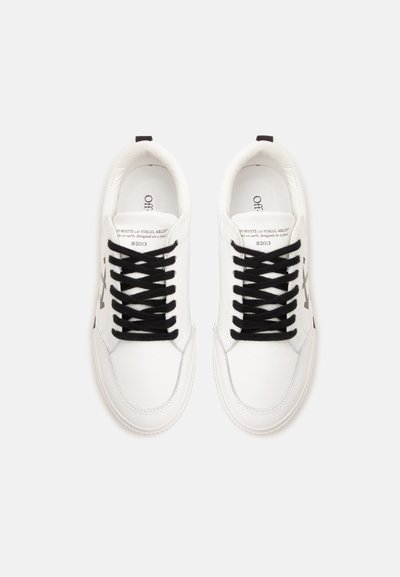Paire de baskets basses blanches avec lacets noirs, détail de texte sur la languette et logo sur les côtés intérieurs, vue de dessus sur fond blanc.
