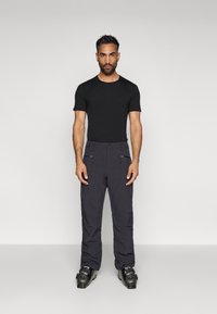 Moški stoji ob enobarvni ozadju in nosi črno prilegajočo T-majico, temne smučarske hlače z zadrgami in črne smučarske čevlje.