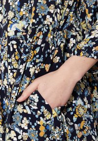 Robe à motifs floraux en bleu marine, ornée de fleurs colorées en bleu, jaune et blanc. Comprend une poche et une taille froncée pour ajouter de la texture.