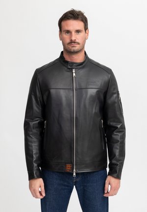Mann trägt eine schwarze Lederjacke mit Reißverschluss und Stehkragen sowie blaue Jeans und steht vor einem schlichten weißen Hintergrund.