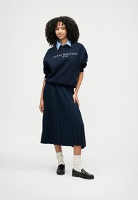 Marinblå sweatshirt med vit "TOMMY HILFIGER"-logotyp, kombinerad med en plisserad marinblå midikjol, krämfärgade strumpor och svarta loafers.