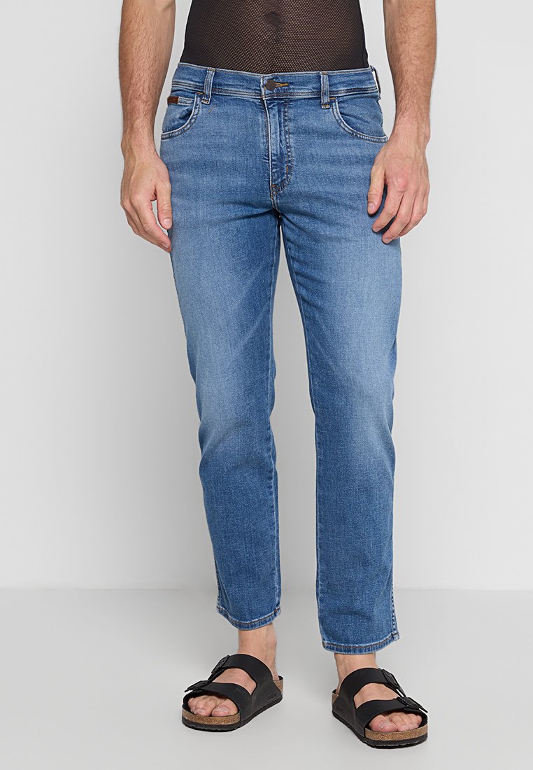 Osoba stoi ubrana w jasnoniebieskie dżinsy i czarne sandały, w siatkowej koszulce, na tle gładkiej białej ściany.