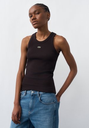 Femeie care poartă un top negru tricotat fără mâneci, cu logo mic verde în formă de crocodil, asortat cu blugi albaștri largi, stând în picioare în fața unui fundal simplu.