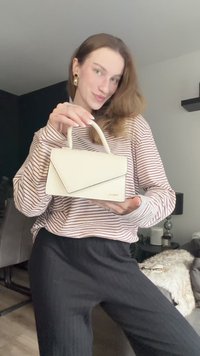 Kleine witte handtas met een gestructureerd ontwerp, met een driehoekige klep en twee korte handvatten, getoond tegen een gestreept shirt met lange mouwen.