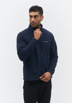 Mann som justerer kragen på en marineblå Columbia fleecejakke, stående mot en ensfarget lys bakgrunn.