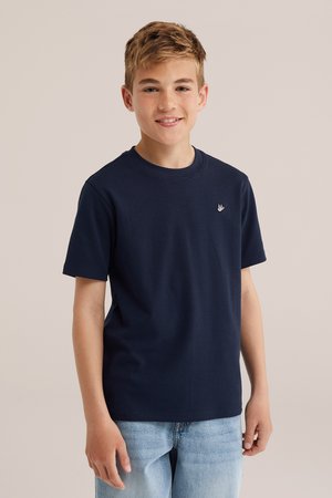 Hymyilevä teini-ikäinen poika, jolla on lyhyet vaaleanruskeat hiukset ja päällään tummansininen lyhythihainen paita sekä vaaleansiniset farkut, yksiväristä taustaa vasten.