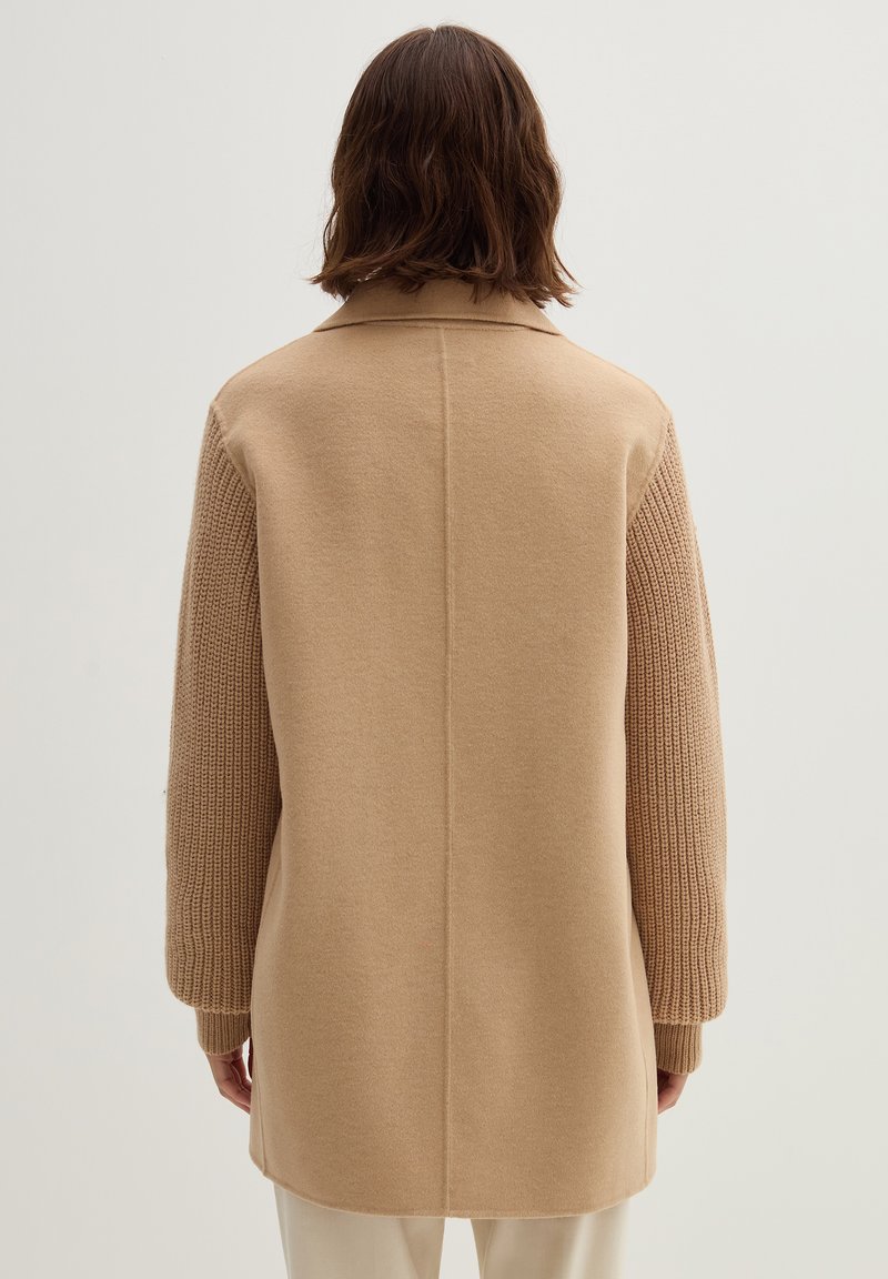 Beige Mantel mit einem Wollkörper und gestrickten Ärmeln, runder Kragen und langer Silhouette, mit gerippten Bündchen und Nahtdetails.