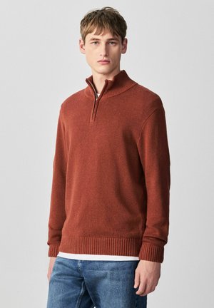 Jaunas vyras, dėvintis rūdžių spalvos megztinį su pusiau užtrauktuku, dėvimą virš baltos marškinių ir mėlynų džinsų, stovintis priešais paprastą šviesiai pilką foną.