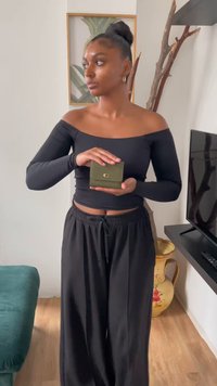 Carteira de couro verde com um fecho dourado, segurada nas mãos de uma pessoa. A pessoa está vestida com uma blusa preta com ombros à mostra e calças pretas.