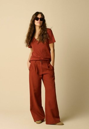 Woman with long wavy hair wears dark sunglasses, rust-red short-sleeve V-neck top, matching wide-leg pants, and beige shoes, hands in pockets.