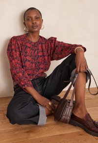 Blusa rossa a motivi floreali con maniche a sbuffo, abbinata a jeans scuri in denim. Accessori includono una borsa a tracolla marrone e mocassini.