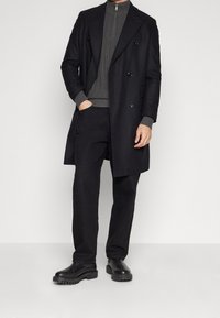 Schwarzer, doppelt knöpfbarer Mantel über einem grauen Rippenpullover, kombiniert mit schwarzen Hosen und klobigen schwarzen Stiefeln. Glattes Material und taillierte Passform.