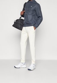 Grå lätt jacka med huva, vita smala byxor och vita löparskor. En svart väska med ett nätfack hålls i ena handen.