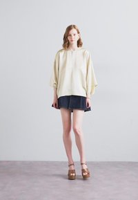 Blouse crème ample avec une fermeture éclair à l'avant, assortie à une mini-jupe en denim et des chaussures à plateformes marron. Texture lisse avec un design décontracté.