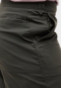 Κοντινό πλάνο σκούρων πράσινων παντελονιών εξωτερικού χώρου με φερμουάρ στην πλαϊνή τσέπη και κεντημένο λογότυπο "Houdini" κοντά στη μέση.