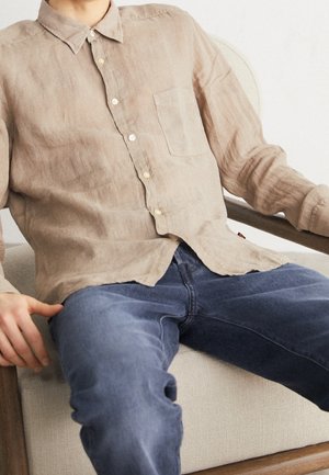 Man draagt beige overhemd met knopen en blauwe jeans, zittend op een stoel met licht stofbekleding en houten armleuningen.