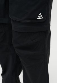 Μαύρο παντελόνι με ενισχυμένες ραφές και τσέπη με καπάκι που φέρει λευκό τριγωνικό λογότυπο ACG στο πλάι.