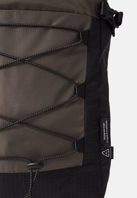 Gros plan d’un sac à dos noir et gris avec des cordons élastiques croisés et une étiquette indiquant qu’il est fabriqué en nylon recyclé.