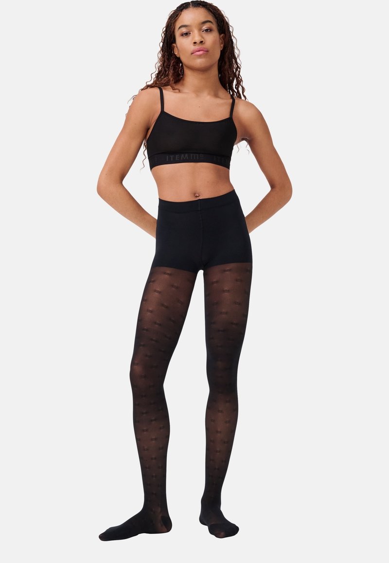 Jeune femme portant un débardeur court noir et des collants noirs transparents à motifs, debout les mains derrière le dos contre un fond uni.