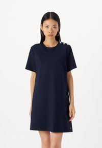Rochie bleumarin cu manecă scurtă dintr-un material neted. Prezintă un guler rotund și patru nasturi aurii pe umărul stâng. Siluetă dreaptă.
