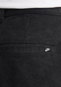 Svarta kordflorsbyxor med fin ribbing, en bakficka med en liten Nike-logotyp. Tyget har en strukturerad yta.