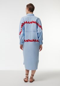 Lichtblauwe jurk met lange mouwen, voorzien van rode zigzag borduursels rond het midden. Glad materiaal met een knie-lange snit.