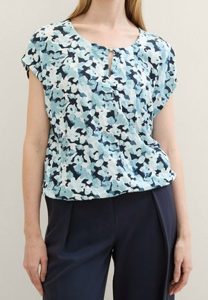 Vrouw die een blauw-witte bloemenblouse met korte mouwen en een kleine keyhole-halslijn draagt, gecombineerd met donkerblauwe op maat gemaakte broek.