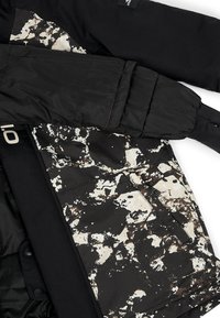 Chaqueta negra acolchada con una tela de patrón abstracto en blanco y marrón texturizada en la parte baja delantera. Cuenta con un bolsillo con cremallera y mangas con puños.