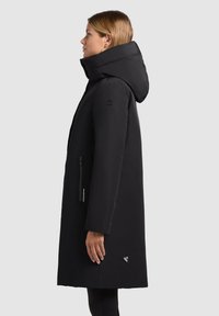 Manteau waterproof noir avec un col haut et une grande capuche, comportant une poche latérale zippée et un patch logo sur l'ourlet.
