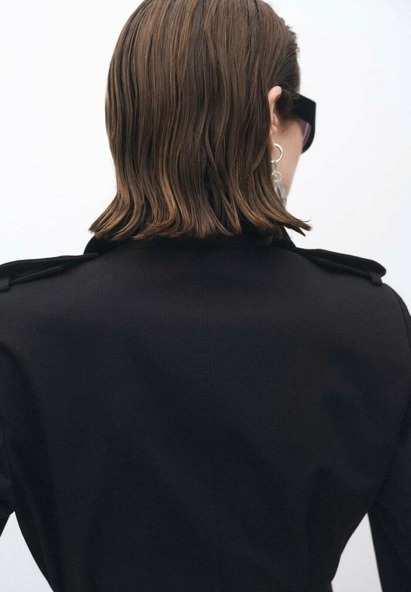 Personne aux cheveux bruns mi-longs portant des lunettes de soleil noires et un manteau noir, vue de dos sur un fond clair.