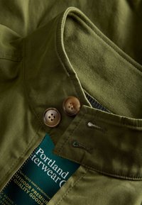 Collar and buttons of an olive green outdoor jacket with visible zipper and partial label reading "Portland Outerwear Co."