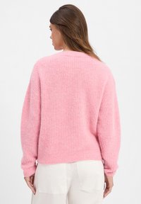 Vrouw met haar rug naar de camera gekeerd, gekleed in een pluizig, roze gebreid sweatshirt en witte broek, tegen een plain witte achtergrond.