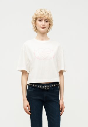 Νεαρό άτομο με κοντά σγουρά ξανθά μαλλιά φοράει λευκό κοντό T-shirt με απαλό τύπωμα καρδιάς και σκούρο τζιν με μαύρη ζώνη με καρφιά.