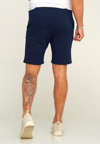 Navy blue cotton shorts with a relaxed fit, hemmed edges, and side seams. Notable tattoo visible on the lower leg. Beige slip-on shoes.